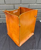 Front and side angled view of the rectangular leather waste basket showing the warm tan color and natural leather texture with metal rivet reinforcements.