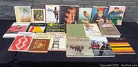 Photo showing 15 softcover coffee table books arranged in two rows on black surface against gray background. Titles visible include Viva Picasso, Van Gogh, Meyer's Ornaments, Complete Guide to French & Canadian Antiques, and others. The books have colorful covers featuring artwork, photographs, and typography.