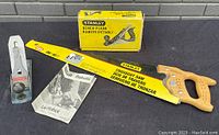 Photo showing Stanley crosscut saw with wooden handle and bright yellow blade, Stanley bench plane, original yellow box, and manual.