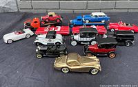 Top-down image showing all 12 diecast toy cars arranged on black cloth with brick wall background. Different models, colors visible including a gold car in front.