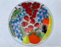 Top view of 14-inch round fused glass charger plate featuring colorful fruit and berry design including strawberries, grapes, cherries, pepper, eggplant, tomato, and banana.