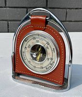 Front and angled side view of vintage horse stirrup barometer and thermometer showing chrome-plated stirrup and brown leather backing with stitched detail and weather dial face.
