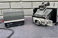 Photo showing front and side angle of Polaroid Sun 600 LMS camera with carrying strap and Polaroid Land Camera Model 95 with attached flashbar holder and lens.