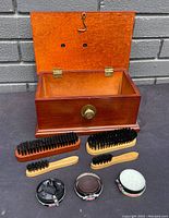 Open wooden shoe shine box showing interior and five shoe brushes in front along with three tins of shoe polish in black, brown, and neutral colors.