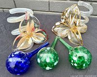 Four art glass watering globes and two clear spiral glass stems with floral accents arranged on dark fabric in front of brick wall.