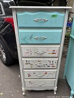 Front and side view of the vintage dresser showing the blue painted top drawers and white lower drawers with stickers and pen marks. Drawer pulls are ornate metal in white.