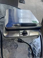 Front and side view of closed stainless steel Cuisinart waffle maker showing handle, temperature control dial, and power indicator lights.