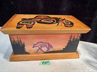 Front and top view of the wooden box showing the main painted Native animal design on the lid and a wolf on the front side with surrounding trees.