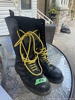 Pair of black Doc Martens Jadon combat leather boots on glass table outdoors showing yellow laces and visible wear.