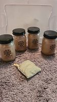 Four small glass jar candles with black lids placed on a carpet along with a small scented sachet pouch in front.