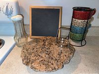 Kitchen items displayed on stove showing granite trivet, salad dressing shaker, soup cup rack with cups, biscuit cutter, and wood-framed chalkboard.