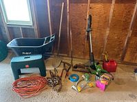 Wide view showing assorted garden tools including shovel, hedge trimmer, grass trimmer, gas can, chain, extension cord, and small tools.