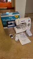 Compact White Mighty Mender W100 sewing machine displayed in front of original box with pedal and instruction manual laid out on floor.