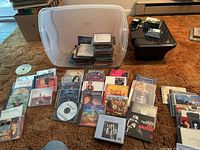 Photo of large plastic bin with multiple stacks and rows of music CDs arranged in and around it, showing numerous CD jewel cases and paper sleeves on orange carpet.