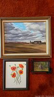 All three framed artworks hung on a wall with orange background: large landscape painting on top, floral painting bottom left, cat photo bottom right.