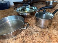 Image showing three stainless steel cookware items on a carpeted surface: a large rounded pan with two handles, a two-handled frypan with glass lid, and a Farberware saucepan with a black handle and glass lid.