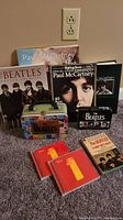 Overview photo showing multiple Beatles books, 2 CDs, and the metal Beatles playing card tin on carpeted floor.