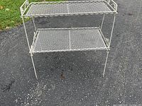 Front and side angle showing two-tier metal plant stand with white lattice shelves and decorative leaf details on the side edges.