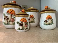 Front view of four white ceramic canisters with textured sides, decorated with multicolored mushrooms and green leaves, topped with brown mushroom cap lids.