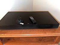 Front view of black ZVOX Sound Bar Sound Base 555 on wooden surface with remote and IR extender cable placed on top.