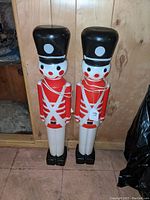 Two 24 inch tall blow mold nutcracker soldier figures standing on floor against wooden wall.