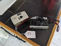 Photo showing both cameras: the black and silver Konex MX-V and the silver Polaroid 2400FF on a dark surface with a white tag marked '53'.
