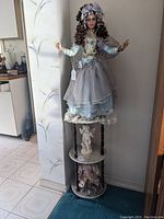Full view of the large doll standing on top of the marble and wood multi-tier stand with cherub figure and two smaller dolls below.