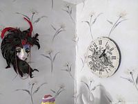 Photo showing the wall mask with peacock feathers and a round clock with Roman numerals and floral design mounted on a wallpapered wall.