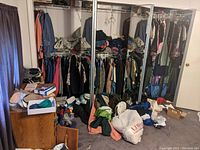 Full view of closet with clothes hanging on racks, folded clothes in shelves, shoes, and scattered clothes on floor.