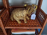 Gold-tone ceramic elephant figurine placed on a woven wood shelf, showing the entire side profile with detailed floral ornamental patterns and golden finish