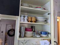 Kitchen cupboard shelves with various stacked plates, bowls, coffee mugs, a cocktail shaker, and a pepper mill.