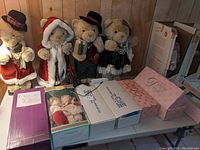 Four teddy bears dressed in Christmas and holiday themed costumes, standing upright and some holding accessories like an accordion, in front of wood paneling. Below them are original boxes for dolls marked with collection names.