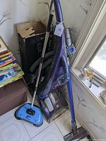 Dyson cordless stick vacuum with blue manual sweeper and Bissell ProHeat 2X partially visible behind, against a wall.