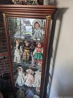 Full frontal view of the glass front curio cabinet containing various porcelain dolls on glass shelves, showing wood frame and glass door with decorative gold accents on corners.