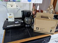 Five kitchen appliance items on black stove: two electric can openers, black drip coffee maker, George Foreman grill, open box for copper crisper indicated.