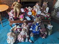 Group of approximately 20 porcelain dolls arranged on and around a wooden coffee table, showing variety in size, color, and style of dolls and their clothing.