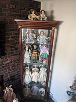 Full view of the glass curio cabinet containing a large group of porcelain dolls in various vintage dresses displayed on multiple shelves, with two dolls sitting on top of the cabinet.