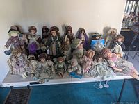 Wide shot of approximately 22 collectible porcelain dolls displayed on a white folding plastic table, showing a variety of vintage-style clothing and hair colors.