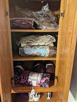 Six throw blankets in wooden shelf: two rolled new Canon fleece throws in pink/black pattern on bottom shelf, four folded used blankets including dark purple, beige knit, patchwork, and taupe on two other shelves.