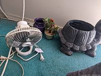 Small white electric tabletop fan with metal blades and round base, power cord and plug visible, placed on carpeted floor.