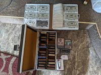 Photo showing a silver carrying case with multiple cassette tapes labeled mostly with artist names, alongside a white plastic binder with cassette tapes inside clear sleeves.