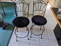 Pair of black wrought iron swivel barstools with upholstered round black seats, shown side by side on tiled floor.