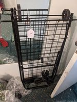 Black folding shopping cart frame leaning against wall, folded flat with visible metal grid panels and stair climbing tri-wheel assembly.