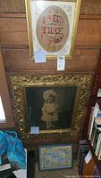 Three framed art pieces displayed on wall and floor: top needlepoint 'I NEED THEE Every Hour', large ornate gilded framed photo of child center, framed 'HAVE A HAPPY DAY' needlepoint on floor below.