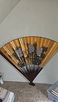 Full view of the large decorative Asian folding fan mounted on the wall, showing the floral painted details on gold background and dark wooden ribs.