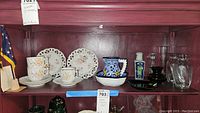 Photo showing shelf display with floral latticework plates, spiral fluted tea cups and saucers, various vases, and the miniature Alaskan flag on the left.