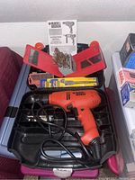 Orange Black & Decker corded drill inside black plastic molded case with assorted drill bits in red plastic holders and instruction manual behind case.