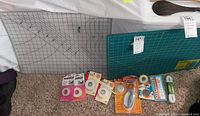 Photo showing two quilting mats (green and clear), Fiskars rotary cutter in package, spare blades, fabric grips, measuring tape, HeatnBond iron-on adhesive, Stitch Witchery fusible bonding web, white fabric pencils, and elastic strips arranged on carpeted floor.