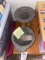 Two basalt molcajetes with their pestles on a wooden surface next to packaging boxes. Set accompanied by handwritten note stating '2 Molcajete Mexican basalt mortar & pestle'.