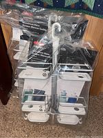 Two boxed Oxo Aluminum Hose Keeper Shower Caddies in original packaging, showing shelves and hooks for holding shower hose.